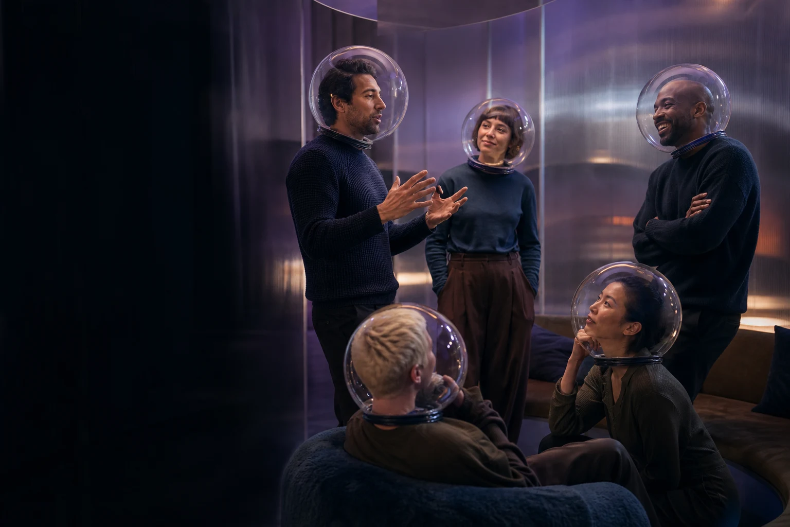 Five people converse in a curved room while wearing identical transparent acoustic bubble helmets